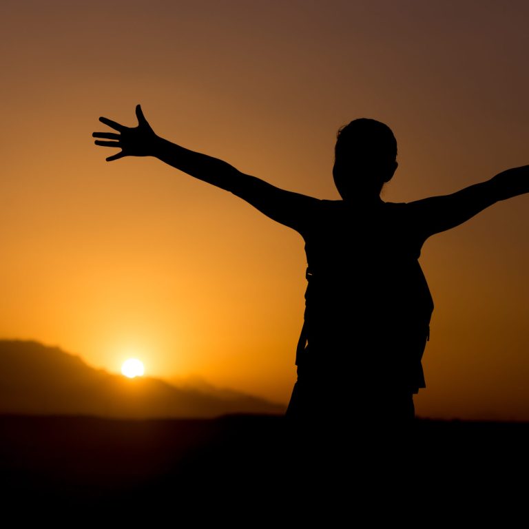 Silhouette of young backpacker person standing with raised arms feeling happy in colorful picturesque landscape, sun rising or setting in mountains on horizon